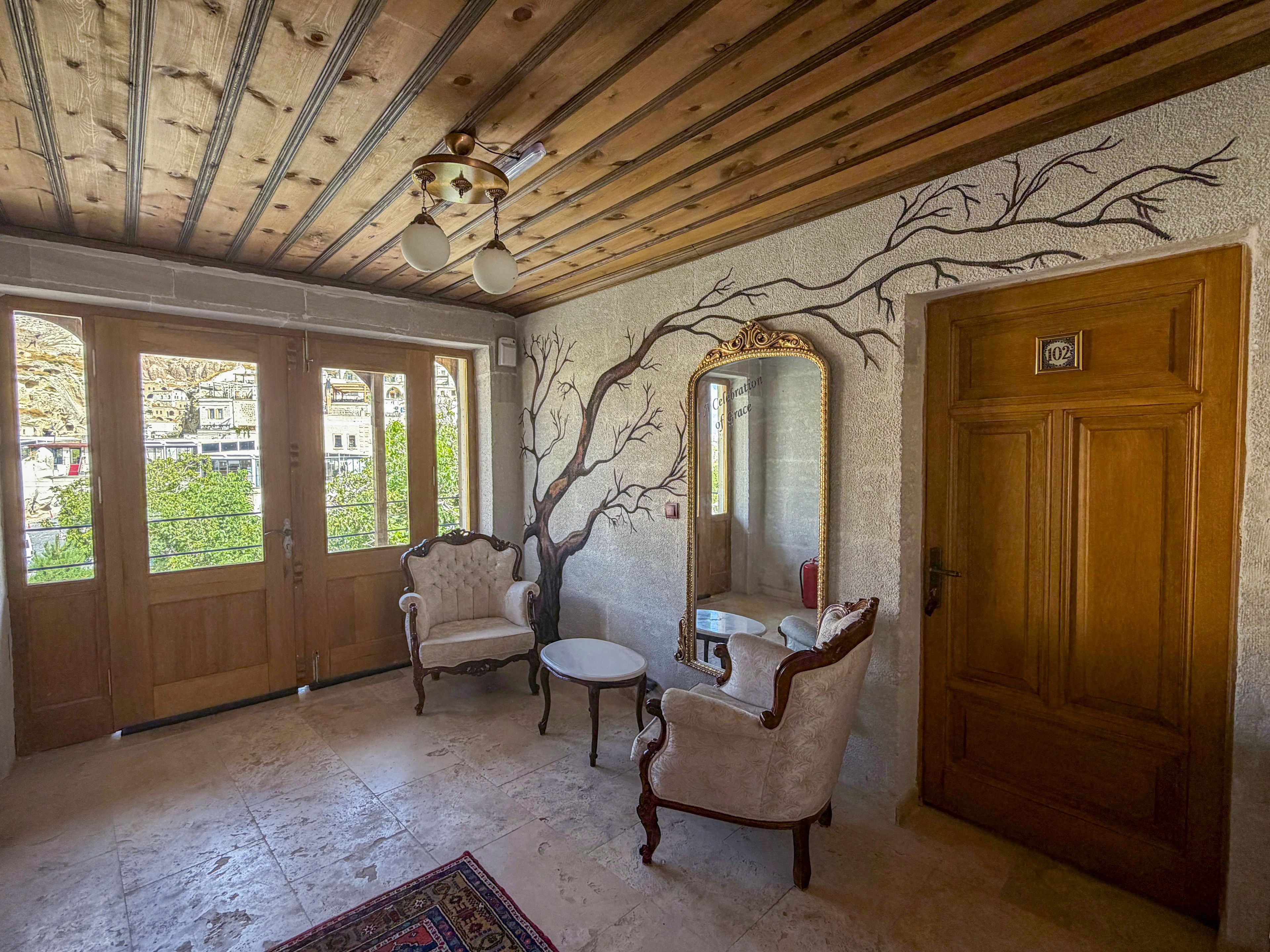 Una habitación con techo de madera, un espejo grande, dos sillas antiguas, una mesa pequeña y una puerta con placa de número.