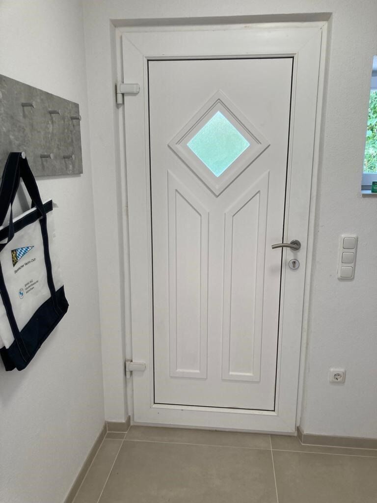 Une porte blanche ornée d’une fenêtre en forme de losange, une plaque fixée au mur et un sol carrelé.