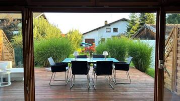 Eine Terrasse im Freien mit Tisch und Stühlen, einem Holzzaun und Blick auf ein Haus und Grünflächen.