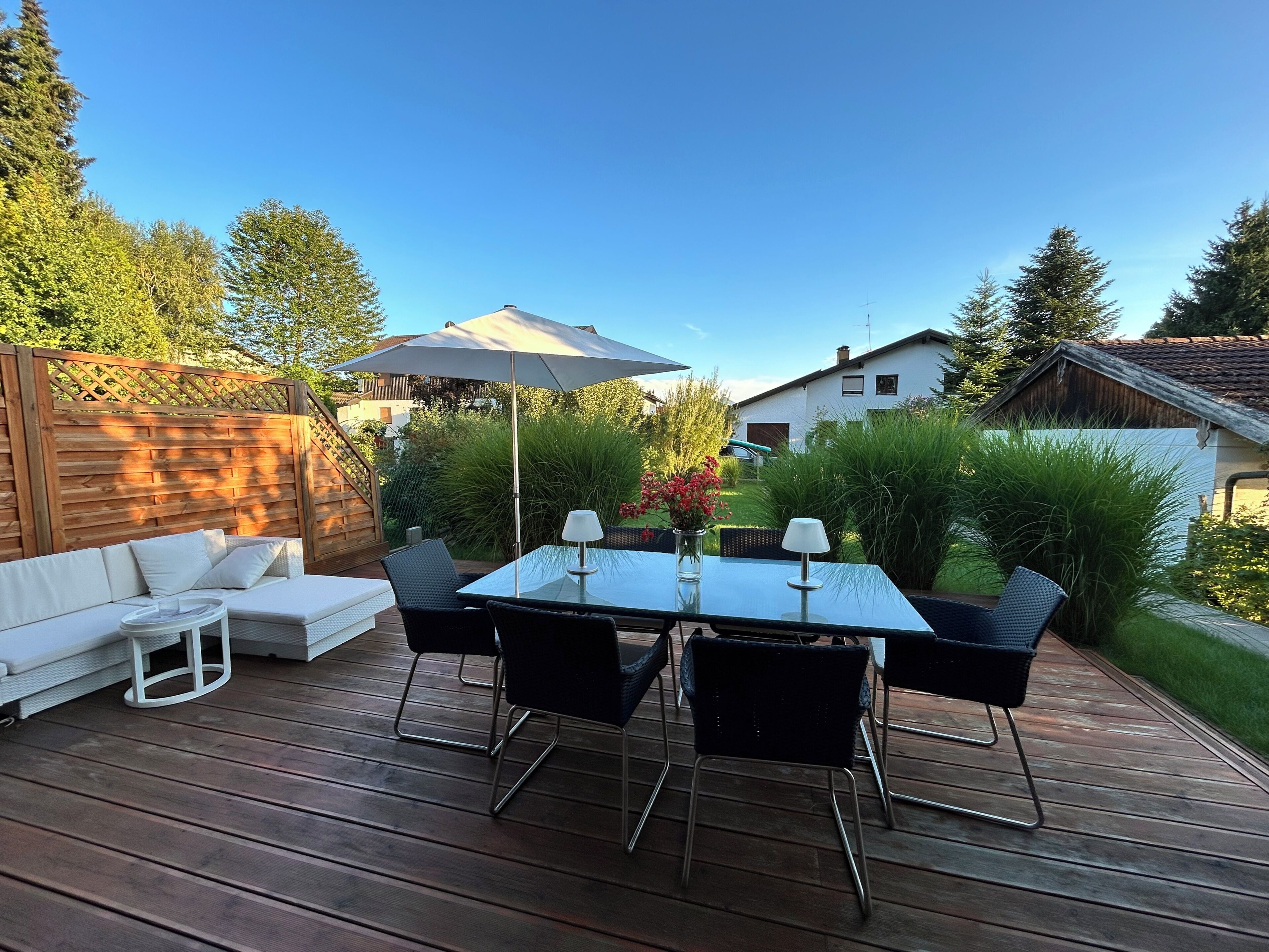 Une terrasse en bois avec une table et des chaises en verre, un canapé blanc et une clôture en bois.