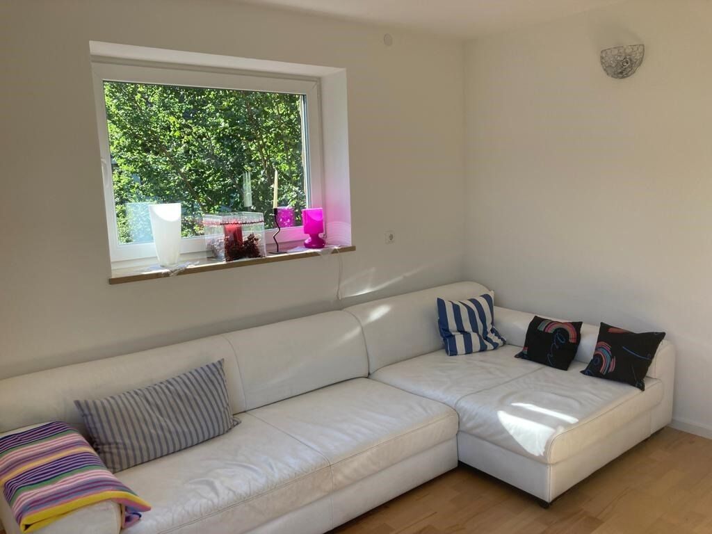 A white sofa with striped and solid pillows, a window with a view of greenery, and a wall-mounted light fixture.