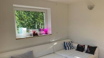 A white sofa with striped and solid pillows, a window with a view of greenery, and a wall-mounted light fixture.