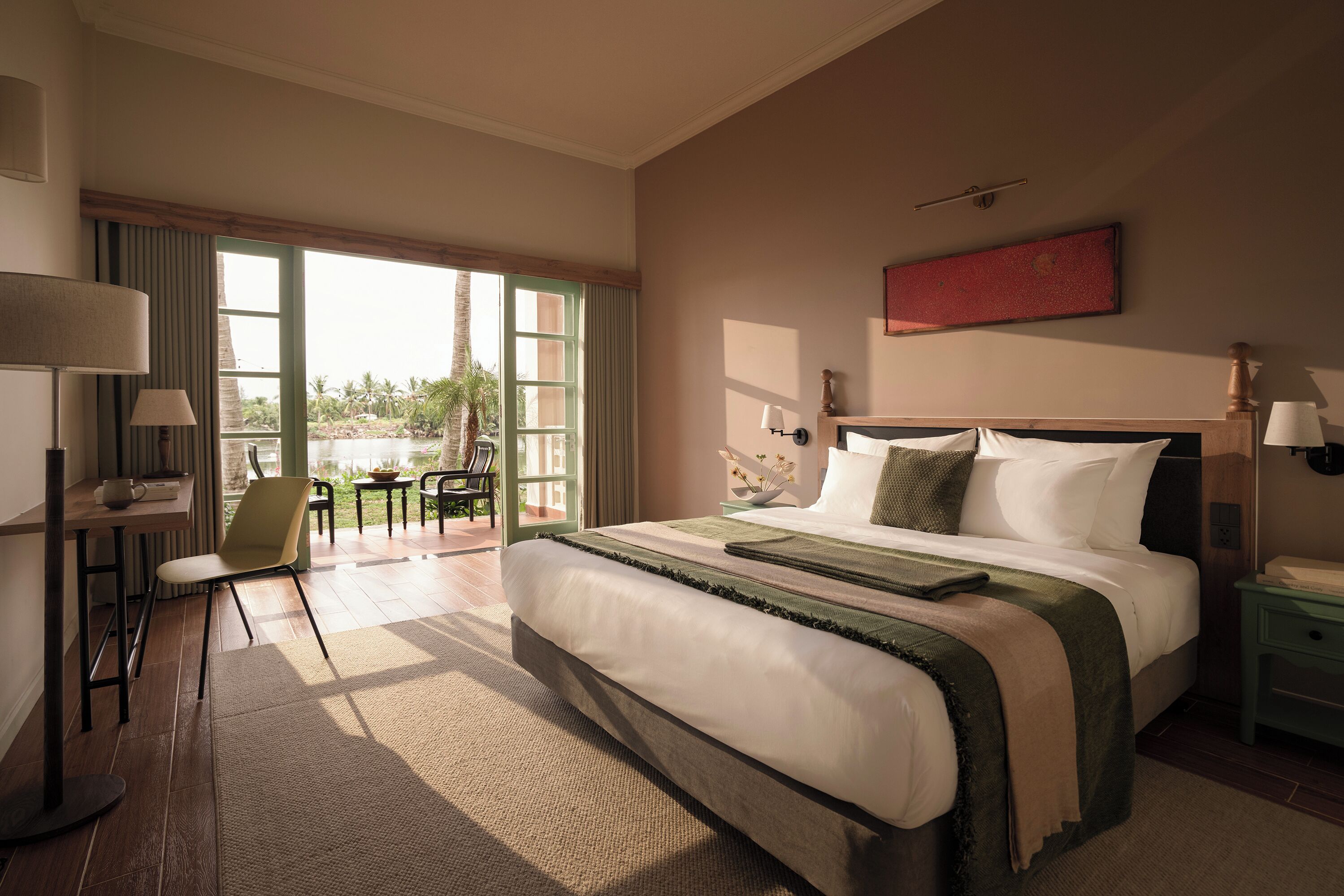 A hotel room with a large bed, a desk with a chair, a window with a view of palm trees, and a red artwork on the wall.