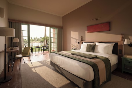 A hotel room with a large bed, a desk with a chair, a window with a view of palm trees, and a red artwork on the wall.