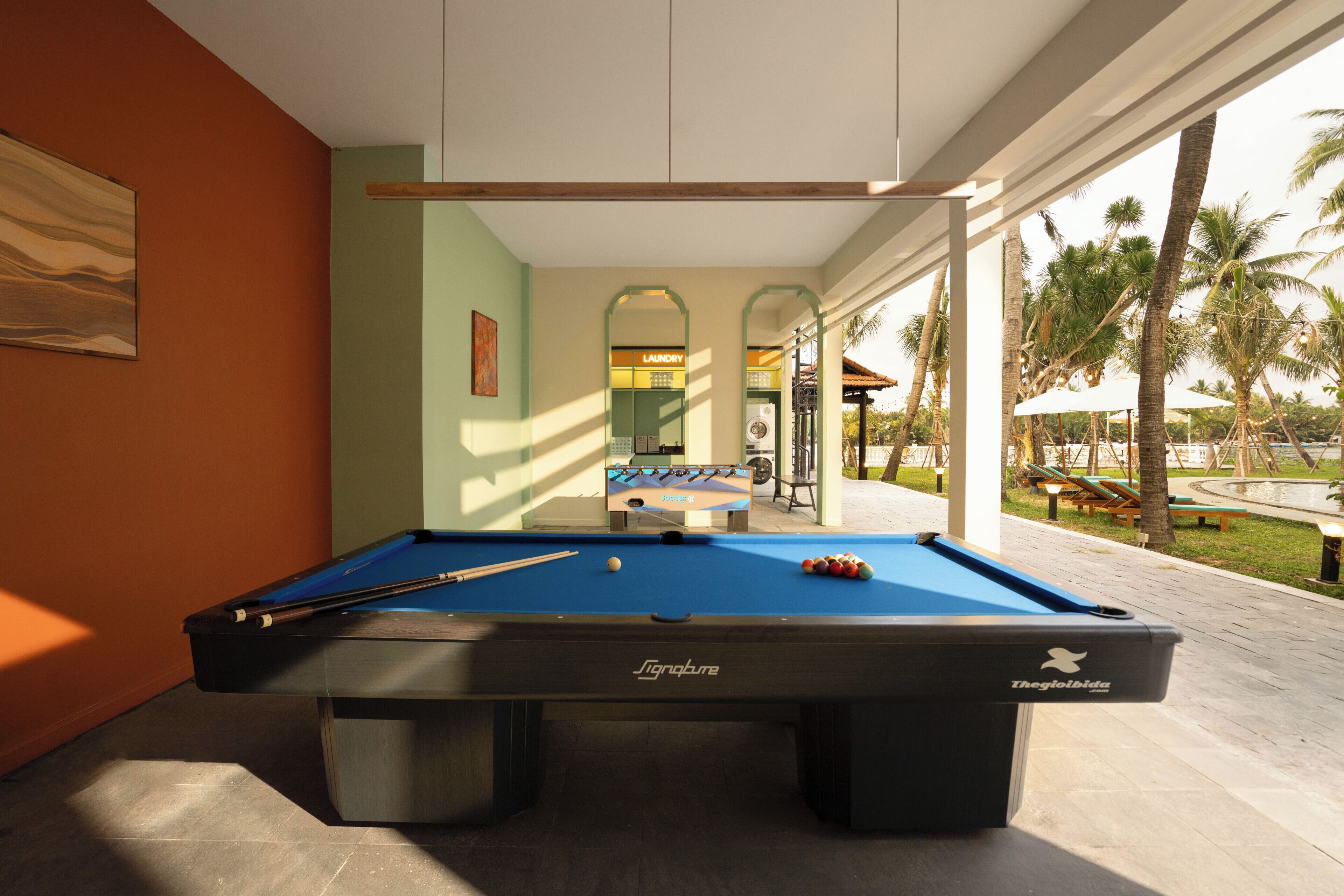 A pool table with a foosball table in the background, a lounge area with palm trees, and a view of a pool.