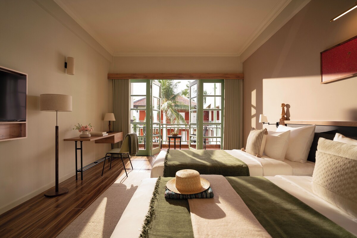 A hotel room with a bed, a desk, a chair, a TV, and a balcony with a view of buildings and trees.