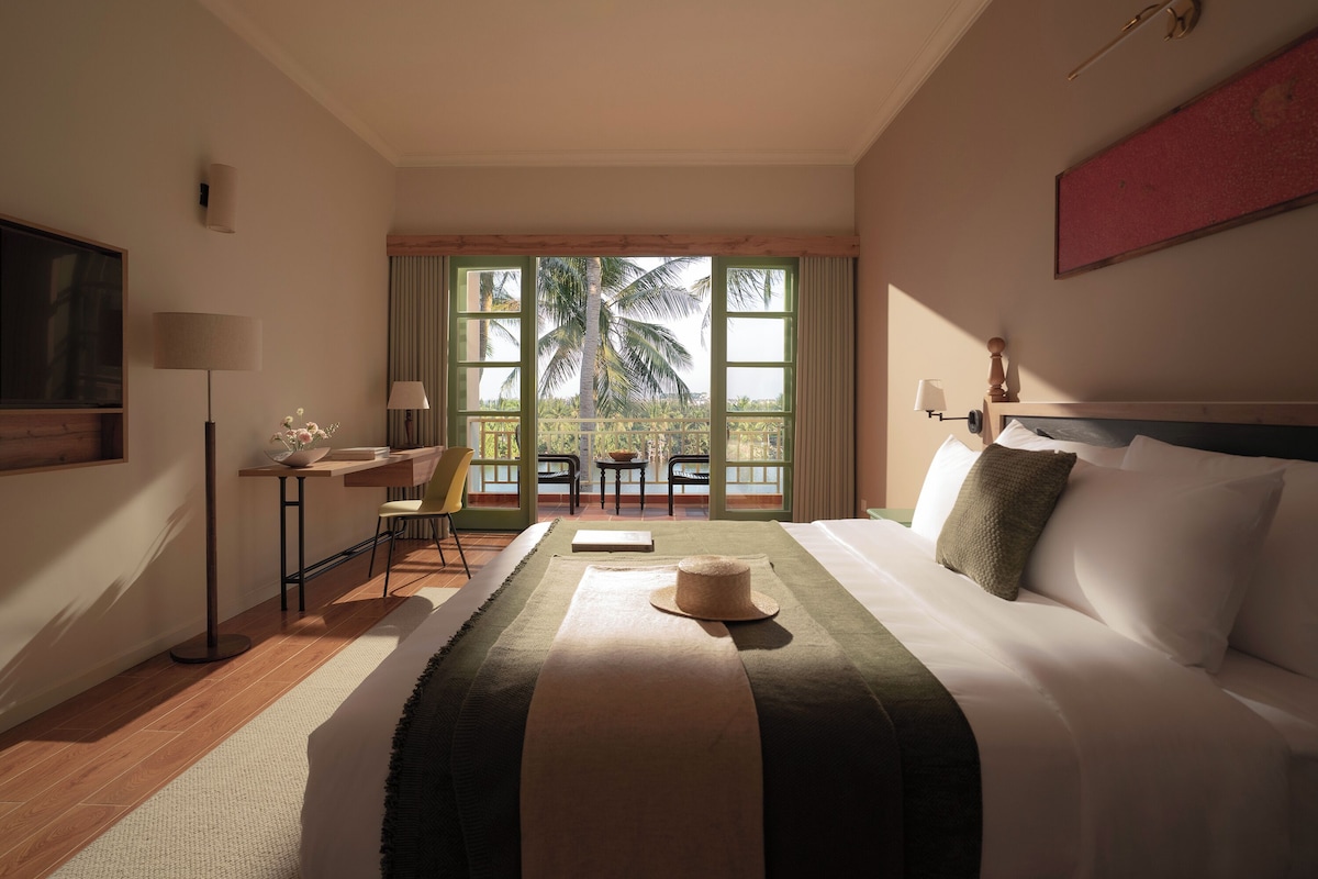 A hotel room with a bed, a desk, a chair, a TV, and a balcony with a view of palm trees.