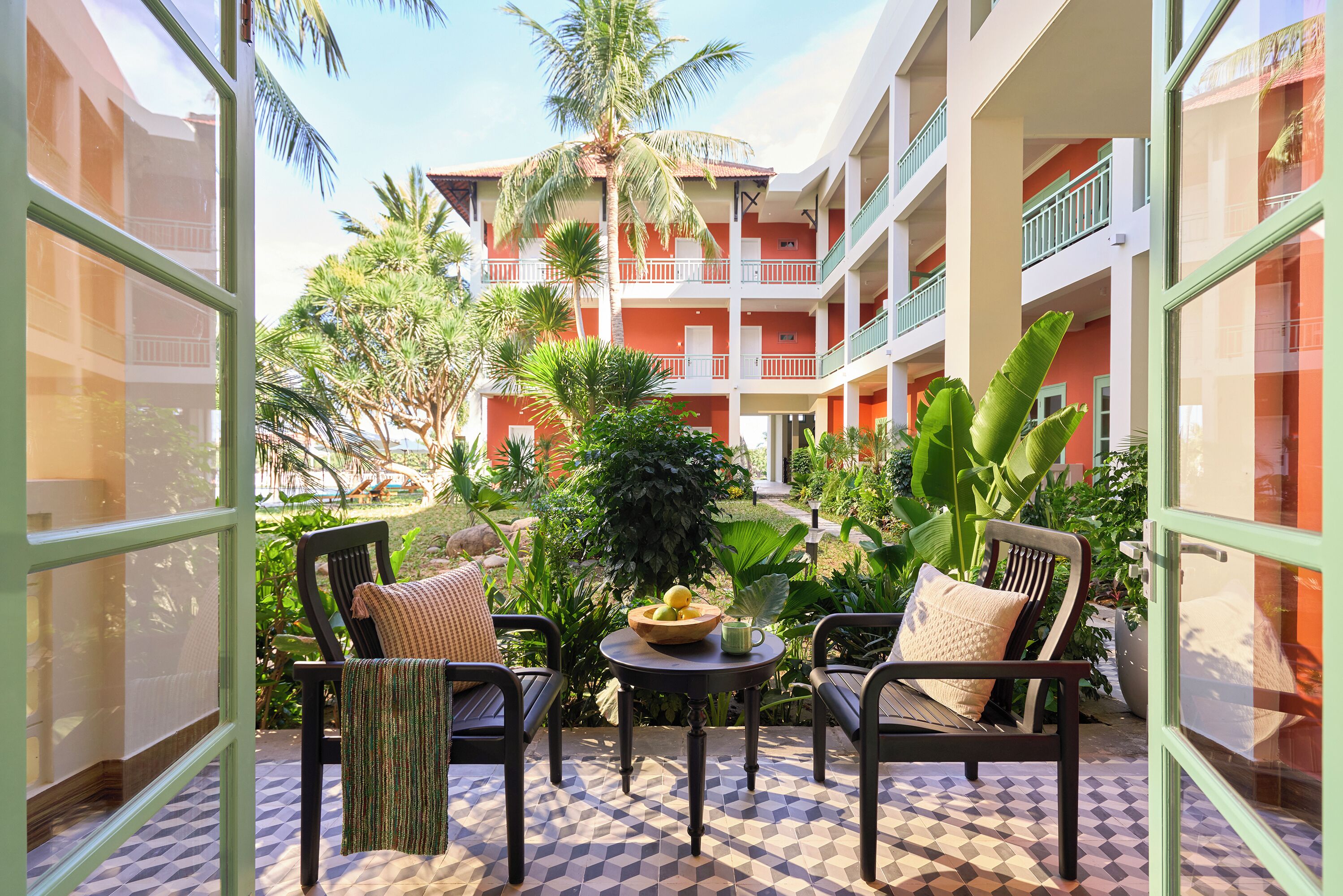 A balcony with two chairs, a small table, and a view of a multi-story building with balconies and palm trees.