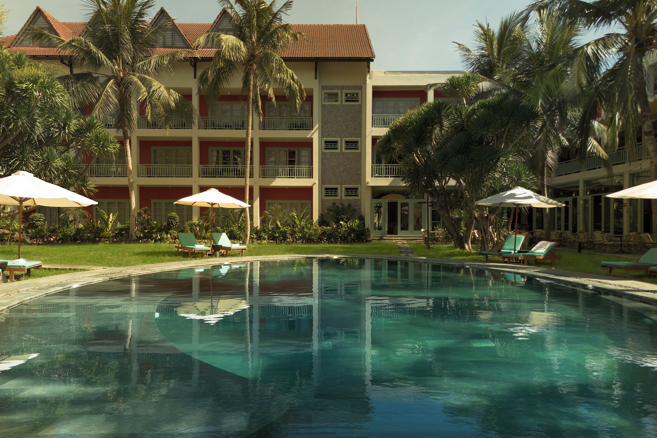 A swimming pool with lounge chairs and umbrellas, surrounded by palm trees and a multi-story building with balconies.