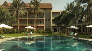 A swimming pool with lounge chairs and umbrellas, surrounded by palm trees and a multi-story building with balconies. - M Village Resort Riverbeach Hoi An (Da Nang)