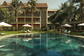 A swimming pool with lounge chairs and umbrellas, surrounded by palm trees and a multi-story building with balconies.