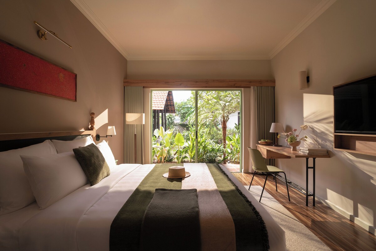 A hotel room with a bed, a desk, a television, and a view of greenery through the window.