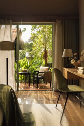 A room with a sliding glass door leading to a garden, a table with a lamp, a chair, and a sofa. - M Village Resort Riverbeach Hoi An (Da Nang)