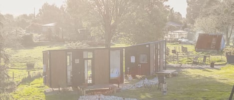 Une petite cabane avec une façade en bois, une porte et une fenêtre. Il y a un espace recouvert de gravier devant la cabane et un arbre à proximité.