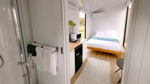 A modern bedroom with a bed, a desk with a computer, a small cabinet, and a shower area. - Rimu View 6 by Tiny Away (Ruatapu)