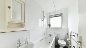 A modern bathroom with a bathtub, sink, toilet, and a window providing natural light. - The Elden - No.16 - Fairway Hosting (Greater London)