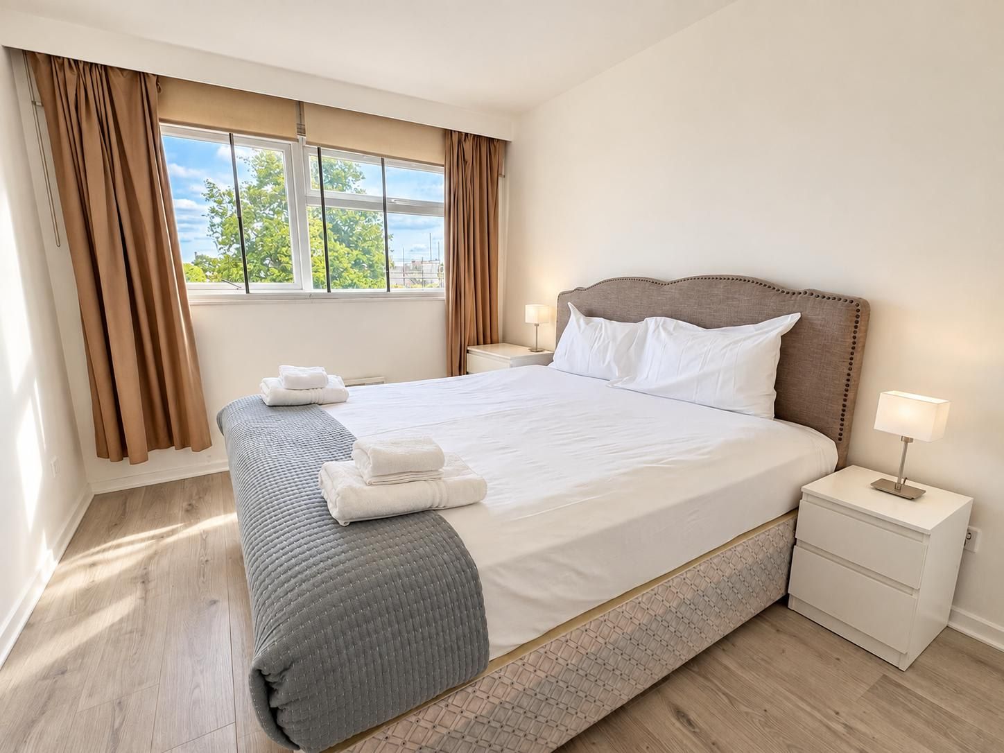A hotel room with a large bed, two bedside tables, a window with curtains, and a view of greenery outside.