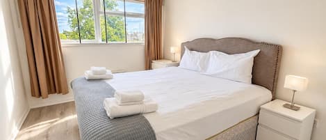 A hotel room with a large bed, two bedside tables, a window with curtains, and a view of greenery outside.