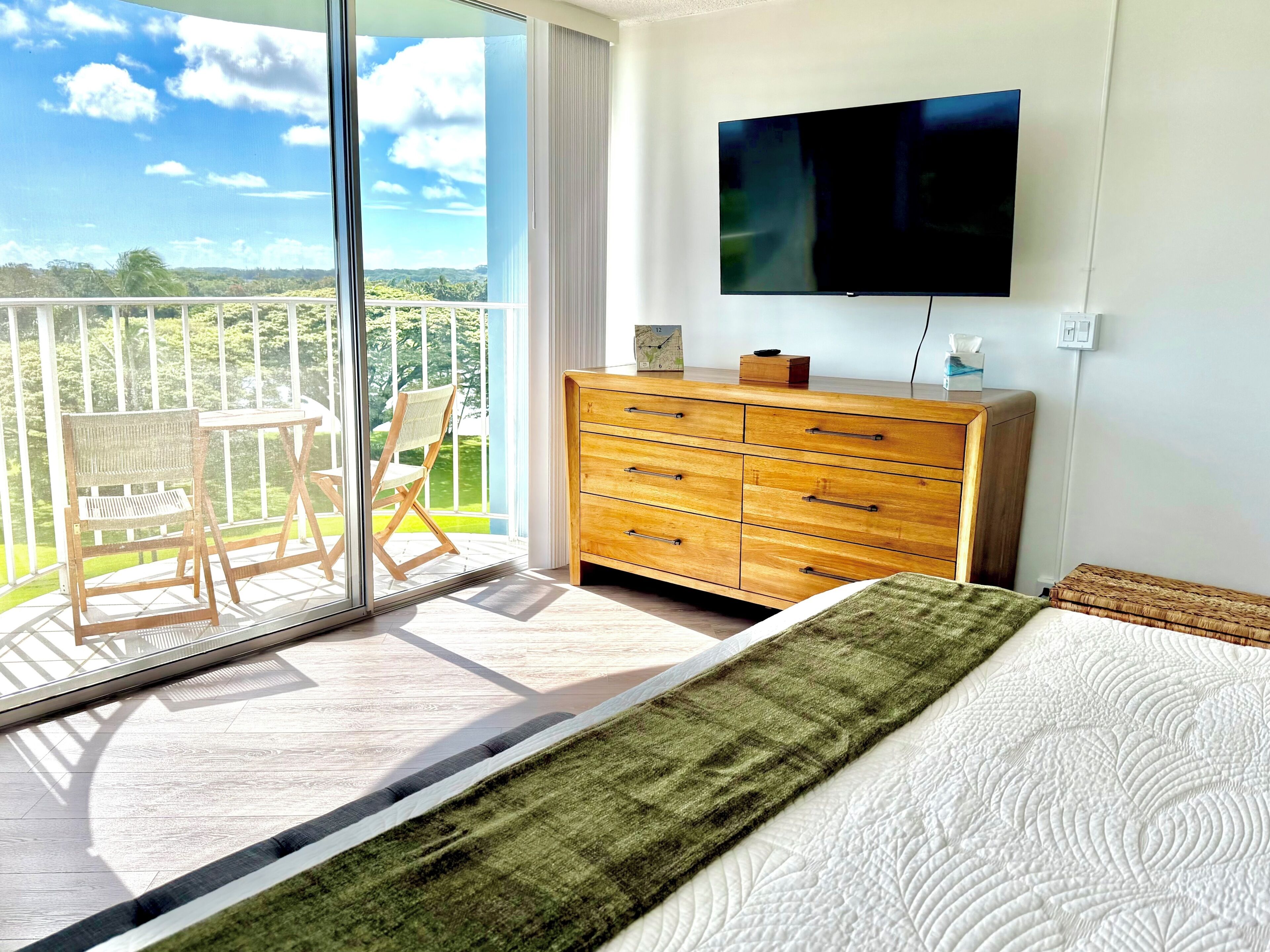 A bedroom with a bed, a wooden dresser, a television, and a balcony with a chair.