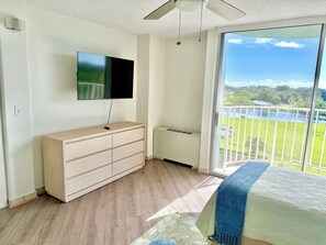 A bedroom with a TV, a radiator, a wooden dresser, a bed with a blue blanket, and a balcony with a view of greenery and water. - Ocean & Wailoa River Views - AC + Pool - 2BD/2BTH (Hilo)