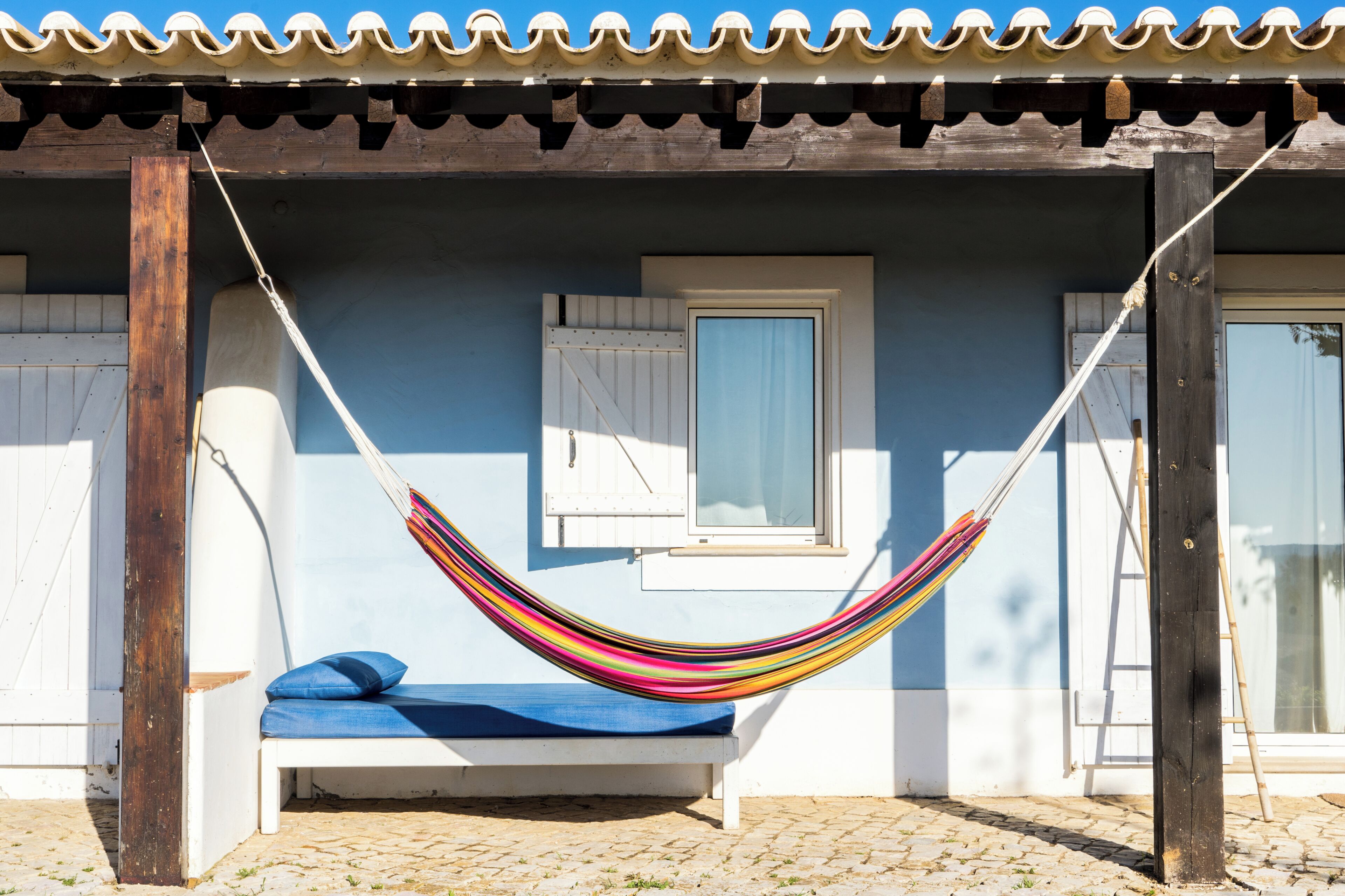 A hammock suspended between wooden posts, a white bench with a blue cushion, and a window with a white shutter.
