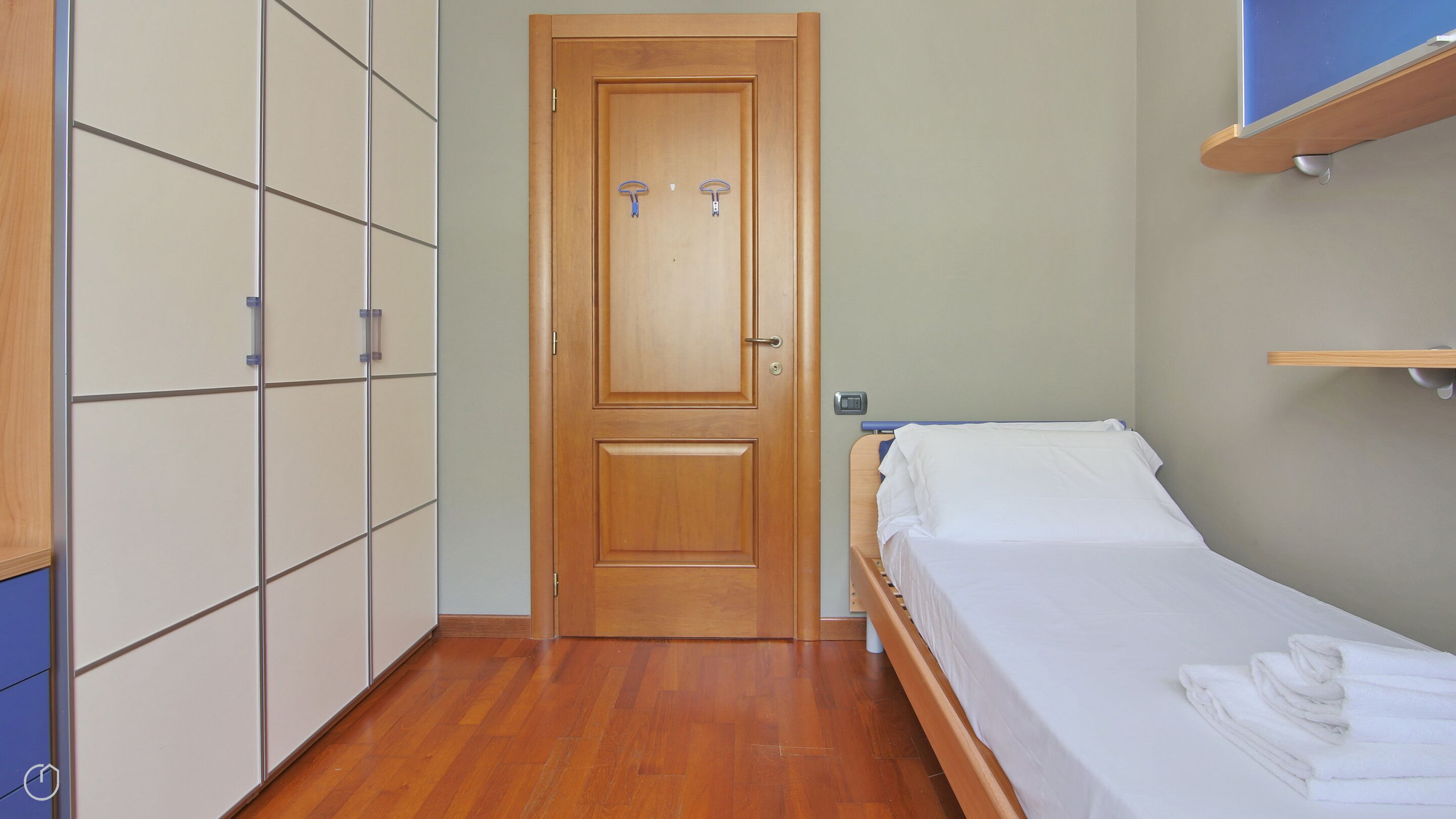 Quarto com porta de madeira, cama com roupa de cama branca, guarda-roupa de madeira e prateleira sobre a cama.