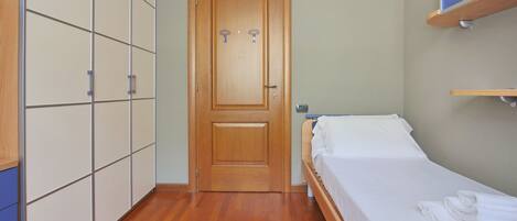 Quarto com porta de madeira, cama com roupa de cama branca, guarda-roupa de madeira e prateleira sobre a cama.