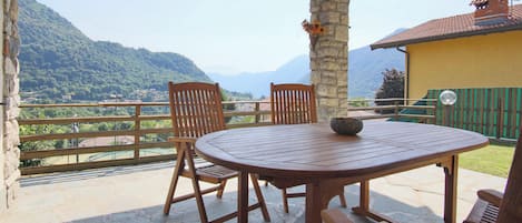Una mesa y sillas de madera en un balcón con piso de piedra, con vista a un paisaje montañoso.