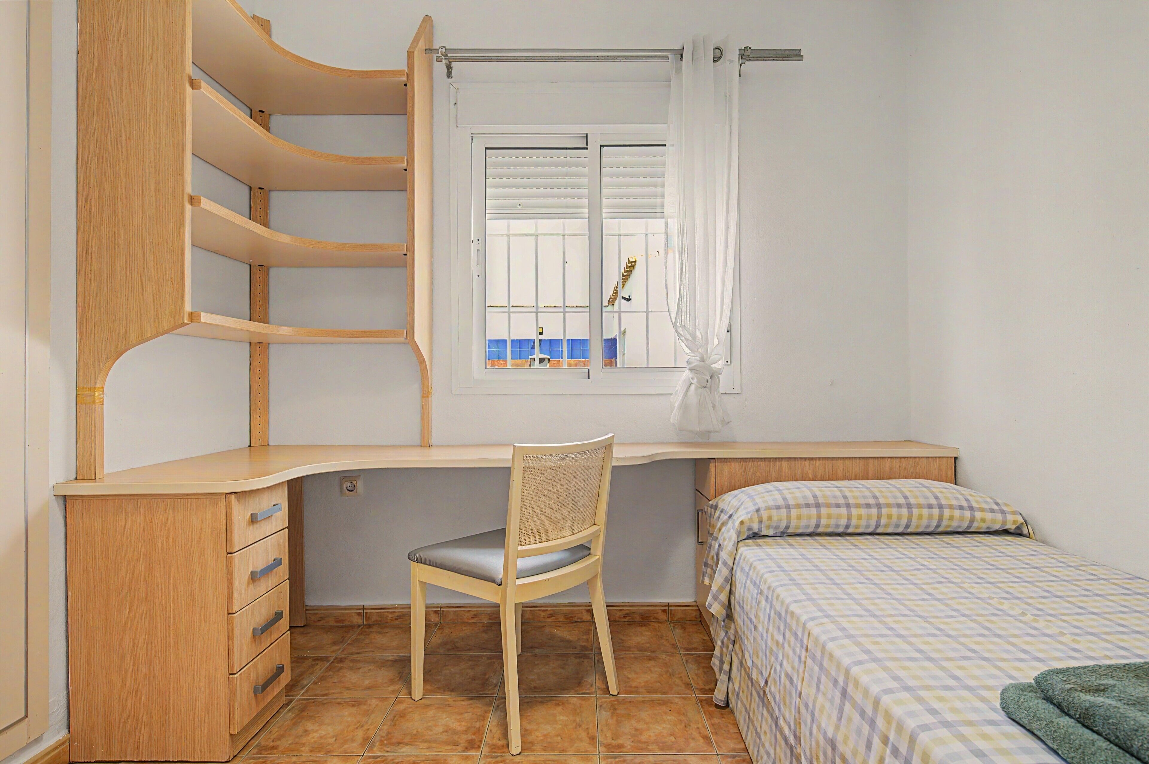 Un dormitorio con un escritorio de madera, una silla, una cama con colcha a cuadros y una ventana con persianas.