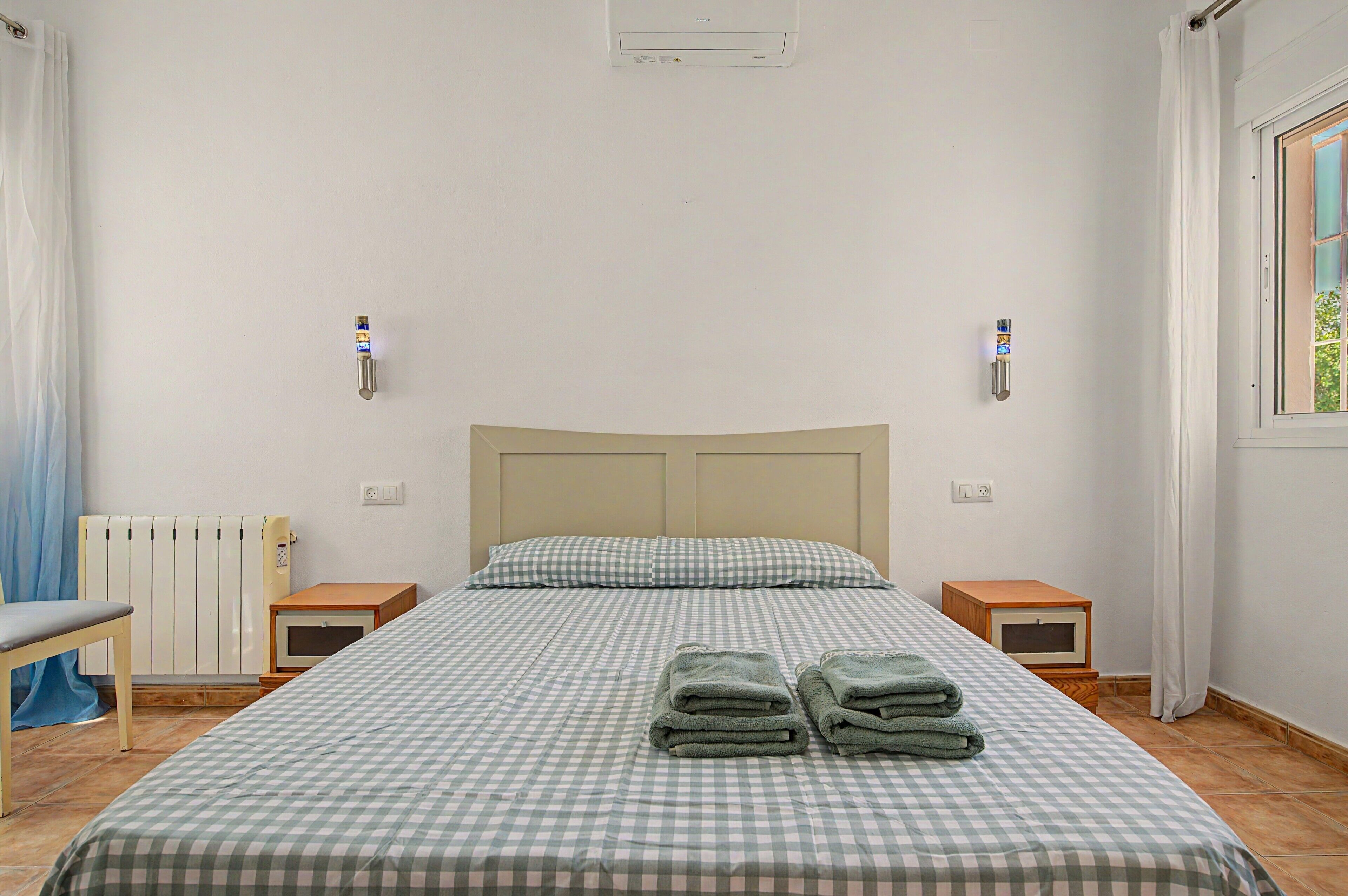 Una habitación de hotel con una colcha a cuadros, dos mesitas de noche, un radiador y una ventana con cortinas.