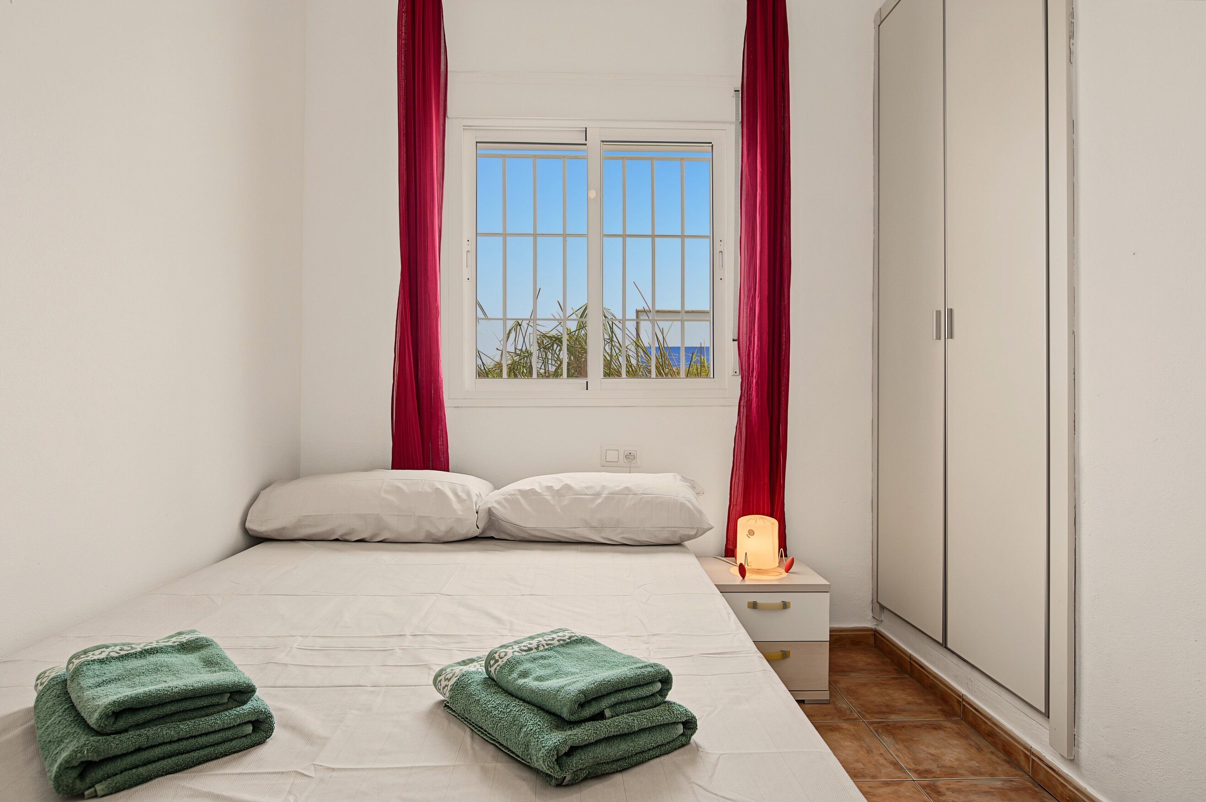 A bedroom with a bed, pillows, a nightstand with a lamp, a window with curtains, and a wardrobe.