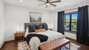 A bedroom with a bed, a ceiling fan, a nightstand, a bench, a window with curtains, and a painting on the wall. - NEW Luxury Cabin | Hot Tub+Wet Bar+EV+Mtn Views (Mineral Bluff)