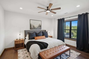 Ein Schlafzimmer mit einem Bett, einem Deckenventilator, einem Nachttisch, einer Bank, einem Fenster mit Vorhängen und einem Bild an der Wand.