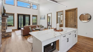 A modern kitchen with a large island, wooden flooring, and a dining area with a sofa and chairs. - NEW Luxury Cabin | Hot Tub+Wet Bar+EV+Mtn Views (Mineral Bluff)