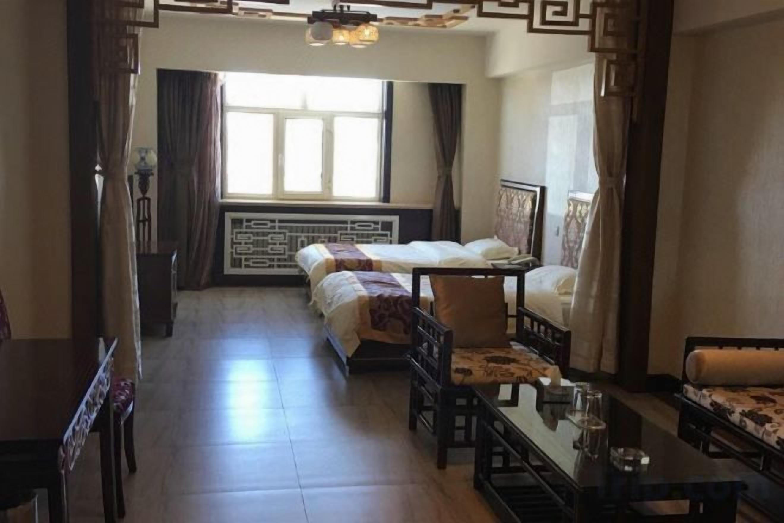A hotel room with a bed, a desk, and a chair. There is a window with curtains, a radiator, and a wall-mounted shelf.