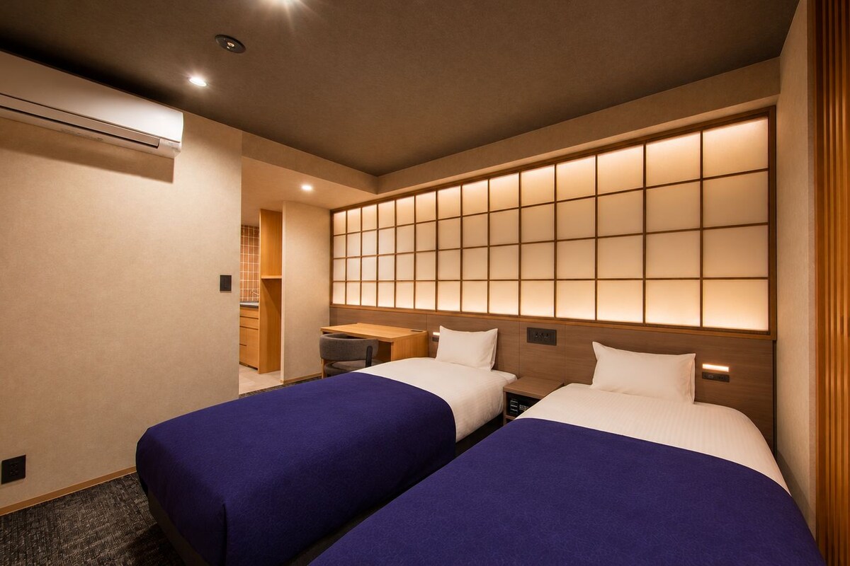 A hotel room with two beds, a desk, a chair, and a wall with a grid pattern.