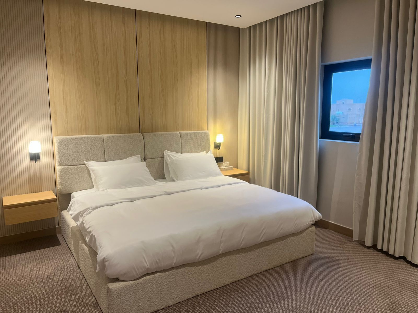 Habitación de hotel con una cama grande, dos mesitas de noche, cabecero y una ventana con cortinas.