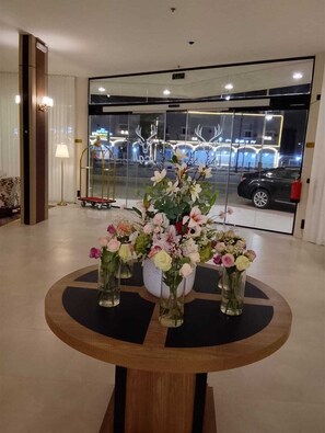 Un vestíbulo con una mesa redonda rodeada de jarrones de flores, una lámpara y una puerta de vidrio que refleja un coche y un edificio.