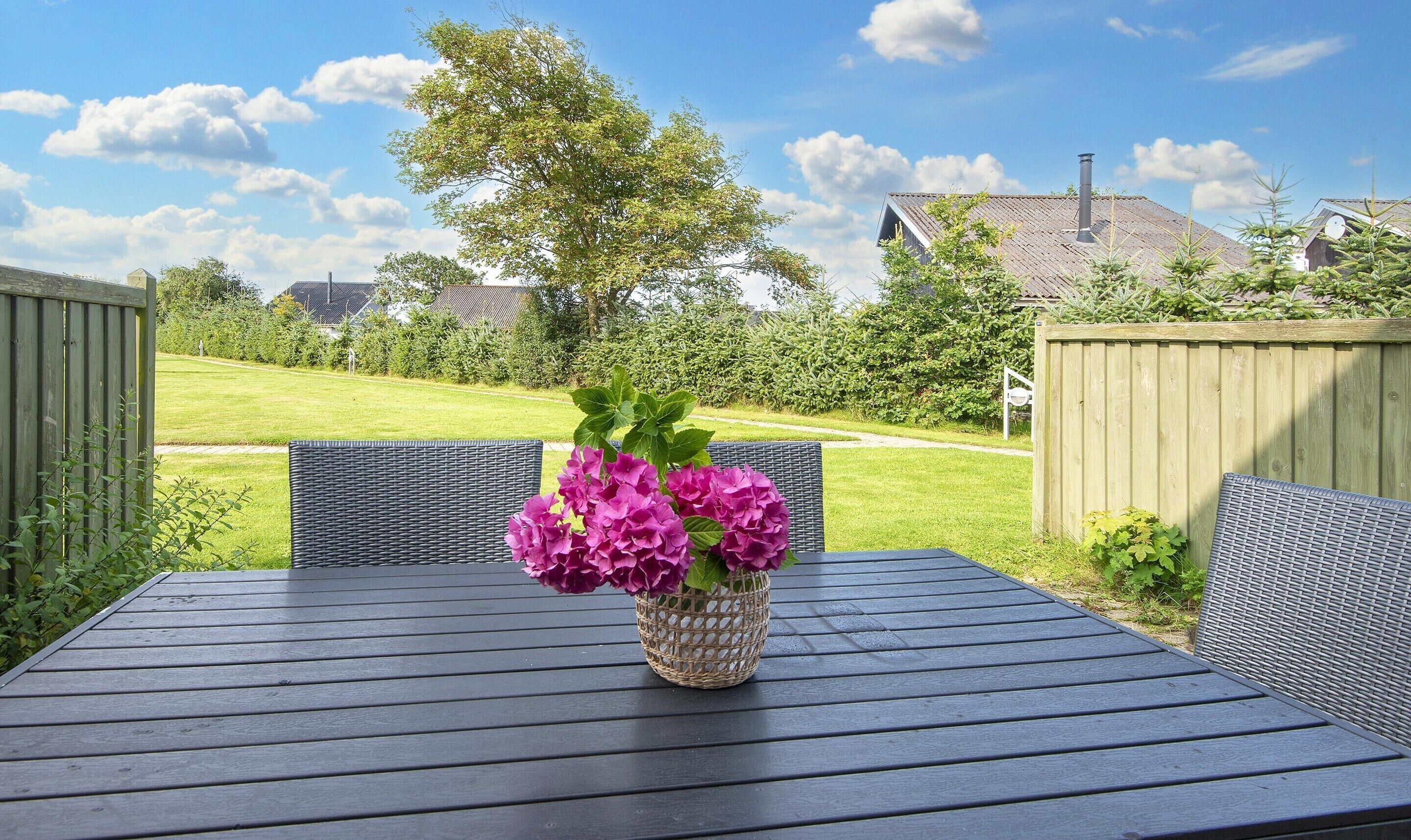 Um pátio externo com uma mesa e cadeiras, um cesto de vime com flores e vista para um gramado e casas ao fundo.