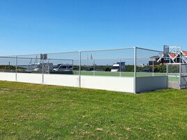 A fenced sports field with a white building and a red-roofed structure in the background.