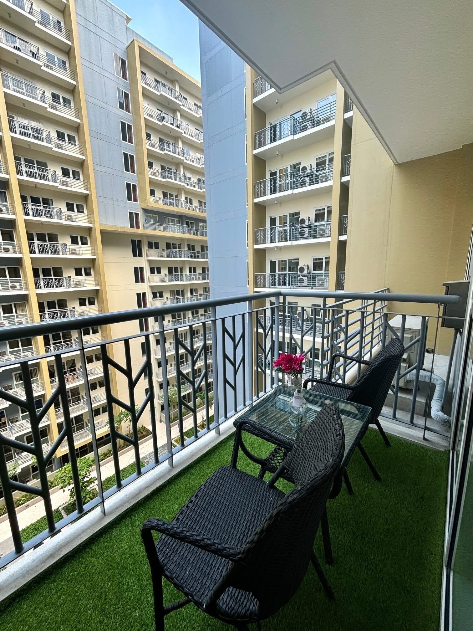 Uma varanda com uma mesa de vidro e duas cadeiras, com vista para um edifício residencial com várias varandas.