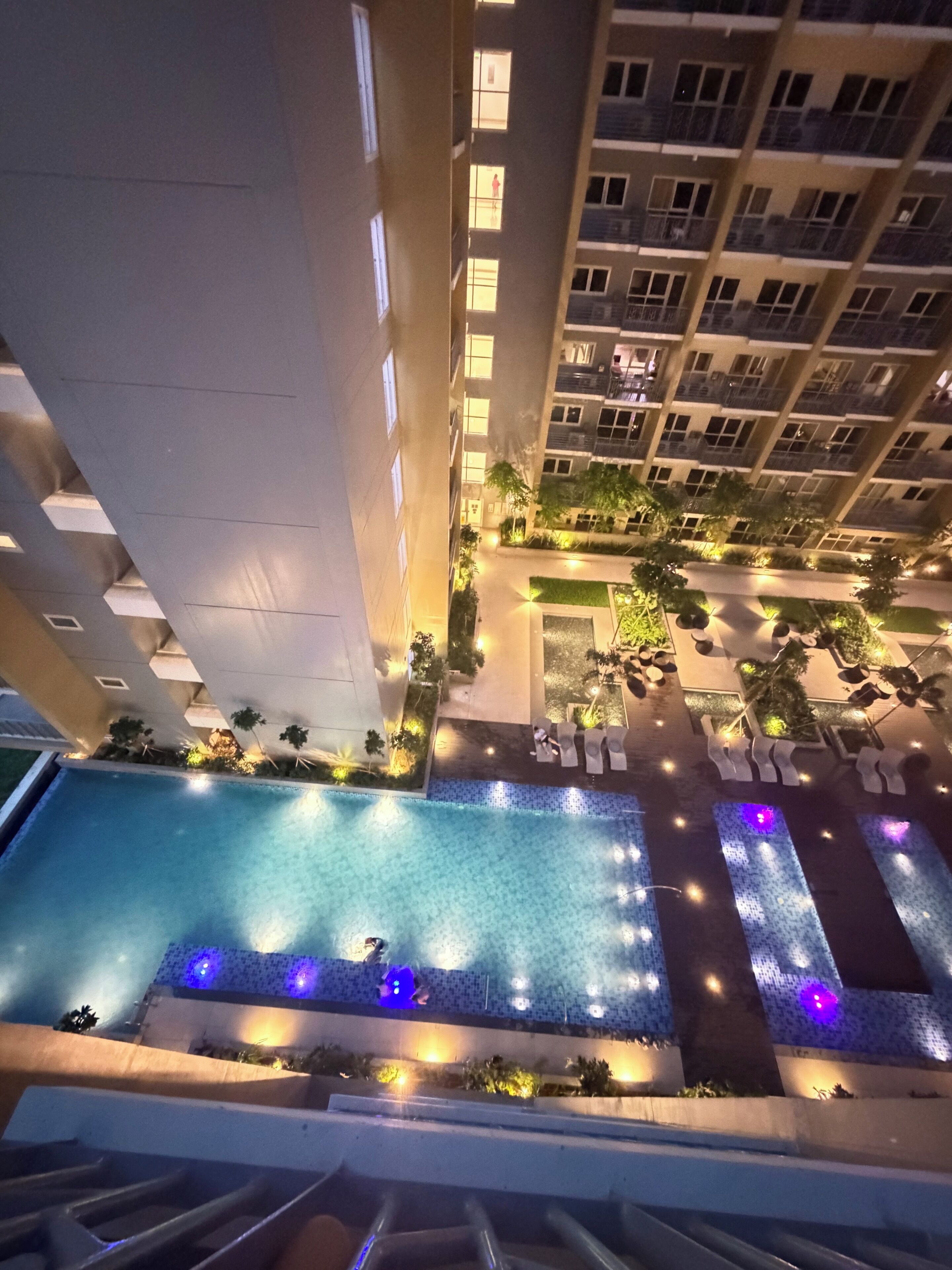 A multi-level residential building with a swimming pool on the ground floor, surrounded by lounge chairs and plants.