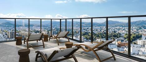 A balcony with four wooden lounge chairs and a small round table, overlooking a cityscape with mountains in the distance.