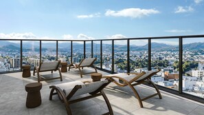 A balcony with four wooden lounge chairs and a small round table, overlooking a cityscape with mountains in the distance. - FAV LUX Asahikawa (Asahikawa)