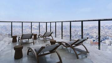 A balcony with four lounge chairs and two small stools, overlooking a snowy mountain cityscape.