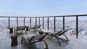A balcony with four lounge chairs and two small stools, overlooking a snowy mountain cityscape.