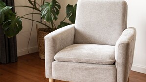 A grey armchair with wooden legs, positioned on a wooden floor, next to a large green plant and a wall-mounted shelf with decorative items.