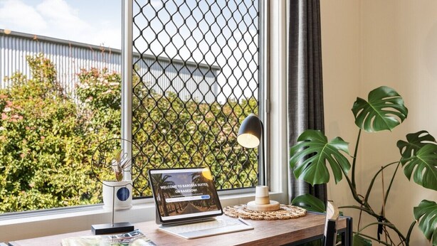 A desk with a laptop, a lamp, and a plant, with a window view of greenery and a metal fence.