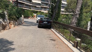 En asfalteret vej med en parkeret bil, en stenvæg og en bygning med flere balkoner i baggrunden.