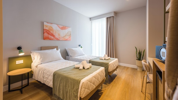 Habitación de hotel con dos camas, un escritorio, una silla, una planta en maceta y una obra de arte colgada en la pared.